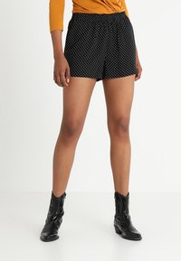 Černé šortky s bílými puntíky, vyrobené z lehkého materiálu. Navrženy s elastickým páskem, doplněné černými kotníkovými botami.