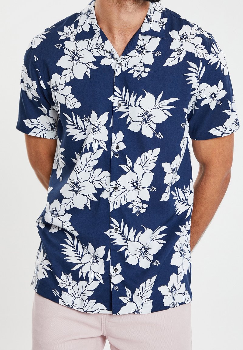 Homme portant une chemise bleu marine à manches courtes avec un grand motif floral blanc et un pantalon clair.