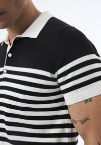 Mustvalge triibuline polo särk, mis on valmistatud puuvillast, kontrastse valge krae ja nööpidega, ribadega varrukateotste ja detailse tekstuuriga.