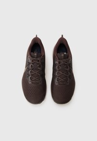 Chaussures de sport marron avec un upper en maille, des lacets plats, un col rembourré et une semelle texturée avec de petites perforations pour la respirabilité.