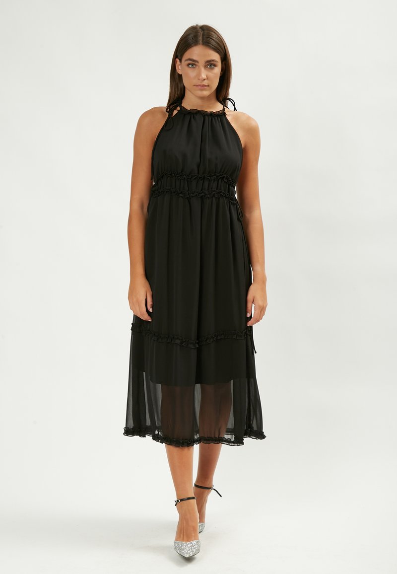 Vestido midi negro sin mangas con un escote y cintura fruncidos, que presenta volantes en las capas y una superposición transparente. Accesorado con tacones plateados.