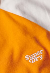 Tela de algodón naranja con una textura suave que presenta un logo bordado en blanco de "Super Dry", sobre un fondo gris claro.