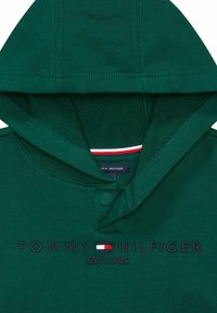 Felpa con cappuccio verde scuro con "Tommy Hilfiger EST. 1985" ricamato in blu navy e un piccolo logo a bandiera rosso, bianco e blu navy tra le parole.