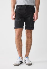 Black denim shorts with a distressed design, featuring frayed edges and rolled hems. Worn with white sneakers and grey t-shirt.