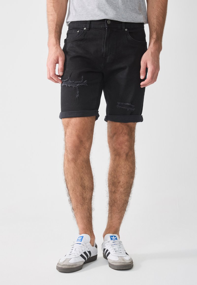 Black denim shorts with a distressed design, featuring frayed edges and rolled hems. Worn with white sneakers and grey t-shirt.