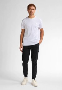 Bílé tričko s kulatým výstřihem a malým logem na prsou, doplněné černými kalhotami typu cargo s bočními kapsami a světlými teniskami.