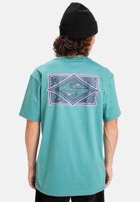 Tyrkysové bavlněné tričko s velkým grafikou na zadní straně v purpurové a černé barvě, se šikmým tvarem a vzorem vln.