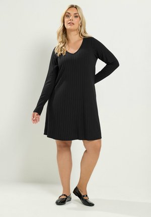 Frau mit langen blonden Haaren, die ein schwarzes, knielanges Kleid mit langen Ärmeln und schwarze Mary-Jane-Schuhe trägt, steht vor einem schlichten Hintergrund.
