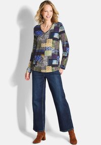 Langärmliges V-Ausschnitt-Oberteil mit einem Patchwork-Design in verschiedenen Mustern und Farben, kombiniert mit dunkelblauen weit geschnittenen Jeans und braunen Schuhen mit Absatz.