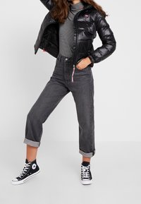 Giacca a vento nera con finitura lucida, maglia a righe sotto, jeans grigi a vita alta e sneakers nere alte.