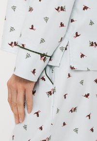 Světle modrá látková košile s červenými a zelenými geometrickými vzory. Má zelené lemování na manžetách a uvolněný střih.