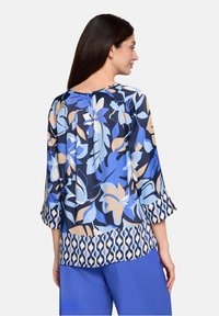 Blouse florale aux tons bleu et beige avec un col rond et des manches 3/4. Présente un dos froncé et un ourlet à motif. Confectionnée en tissu léger.