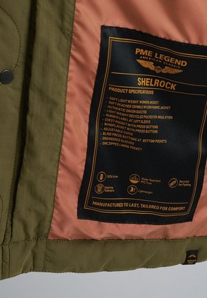 Olivgrüne Nylon-Arbeitsjacke mit einem pfirsichfarbenen Futter; ausgestattet mit einem Produktmerkmaletikett in Schwarz und Orange, mehreren Taschen und verstellbaren Bündchen.