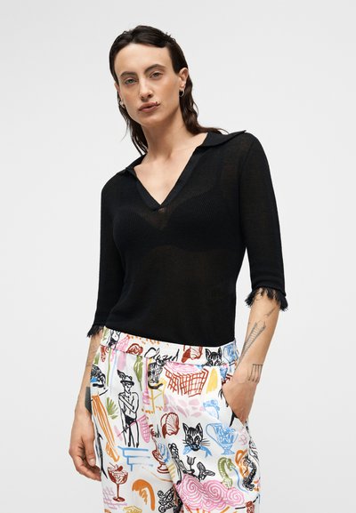 Μαύρο πλεκτό πουλόβερ με V-λαιμόκοψη και κοντά μανίκια με τσαλακωμένες άκρες, συνδυασμένο με πολύχρωμο παντελόνι με σχέδια που απεικονίζουν διάφορα έργα τέχνης.