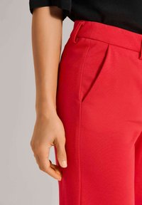 Rote Hose aus glattem Stoff, mit gefaltetem Vorderteil, Gürtelschlaufen und ordentlich verarbeiteten Taschen. Eine Hand hält die Hose seitlich.