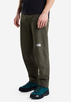 Man draagt olijfgroene The North Face broek en turquoise wandelschoenen met zwarte zolen op een witte achtergrond.