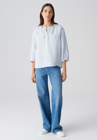 Lichtblauwe linnen blouse met ronde hals en strikdetail, gecombineerd met lichtblauwe wijde jeans. Model draagt witte sneakers.
