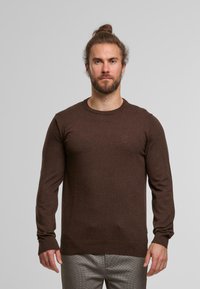 Hnědý svetr s dlouhými rukávy vyrobený z měkkého materiálu, s designem kulatého výstřihu a žebrovanými manžetami. Nosený s vzorovanými kalhotami.