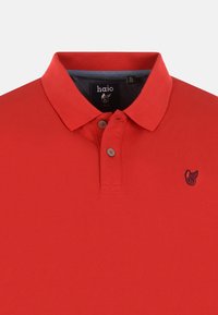 Röd polo-tröja i strukturerad tyg. Har en klassisk krage, treknappslå och en liten marinblå logotyp på vänster bröst.