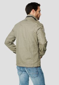 Kaki bunda se střihem do přímého tvaru, měkkou texturou a nášivkami na ramenou. Má vysoký límec a zapínání na zip. Nošena přes modré džíny.