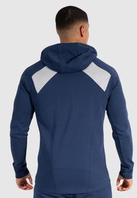 Sudadera con capucha de color azul marino y mangas largas, que cuenta con paneles de malla gris en los hombros y un acabado de tela texturizada. El diseño es ajustado.