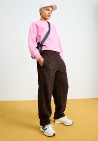 Rosa Sweatshirt, schwarze Sweatpants mit Leopardenmuster, weiße Sneakers mit marineblauen Akzenten und eine weiße Mütze. Das Model steht auf einem gelben Boden.