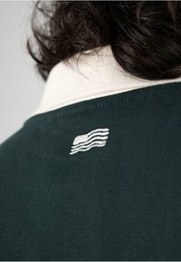 Polo de color verde oscuro con un cuello blanco. Incluye un pequeño logotipo bordado de una ola blanca en la parte posterior.