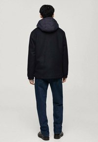 Svart vadderad jacka med grå huva, med en rak passform, bärs tillsammans med mörkblå jeans och svarta skor. Bakvy visas.