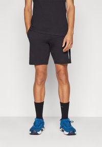 Pantalones cortos de algodón negros con un corte recto, que presentan un sutil detalle del logo en el costado. Combinados con zapatillas azules y calcetines negros.