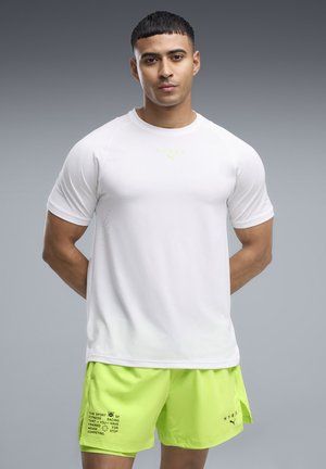 Jeune homme debout, mains dans le dos, portant un t-shirt de sport blanc à manches courtes et un short de sport jaune vif sur un fond gris uni.