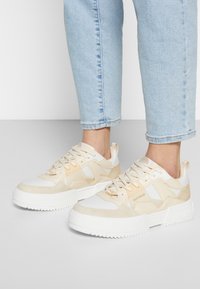 Beiga och vita sneakers med texturerade paneler, platta snören och en kraftig vit gummisula. Logotyp synlig på sidan och tungan.