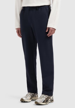 Person trägt marineblaue gerade geschnittene Hose, weißes langärmeliges Hemd und silbergraue Sportschuhe und steht vor einem einfarbigen Hintergrund.