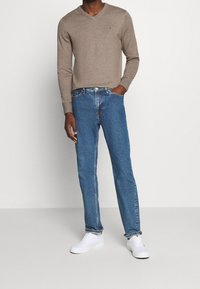 Brauner V-Ausschnitt-Pullover, blaue Jeans, weiße Sneaker. Der Pullover ist gestrickt; die Jeans sind gerade geschnitten mit einer ausgebleichten Waschung und verfügen über fünf Taschen.