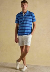 Blau- und marineblau gestreiftes Poloshirt mit einem weißen Kragen, kombiniert mit weißen Shorts und hellen Sneakers, vor einem neutralen Hintergrund.