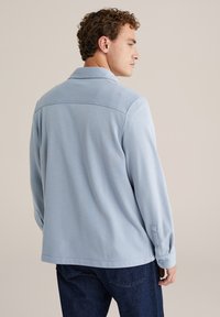 Cămașă albastră deschisă cu guler, mâneci lungi, țesătură texturată și manșete cu buton, purtată cu blugi negri.