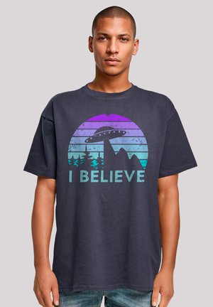 Navy blue cotton t-shirt featuring a graphic design of a UFO above mountains, with the text "I BELIEVE" in distressed aqua letters.