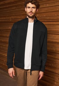 Sort zip-up sweatshirt med et tekstureret design på forsiden, ensfarvede ærmer, høj krave og ribbede håndled, båret over en hvid t-shirt.