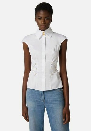 Donna che indossa una camicia bianca aderente senza maniche con colletto alto e jeans azzurri su uno sfondo semplice.