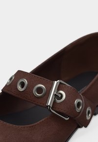 Chaussure Mary Jane en daim marron avec de grands œillets argentés et une boucle métallique argentée sur la bride supérieure.