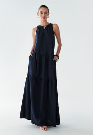 Navy blue sleeveless maxi dress with tiered design, keyhole neckline, side pockets, and flowy texture. Worn with minimal accessories.