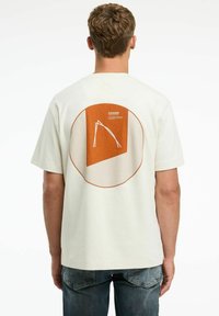 Beige pamut póló rövid ujjakkal, hátán geometrikus narancssárga és fehér mintával, valamint egy textúrált kör alakú mintázattal.