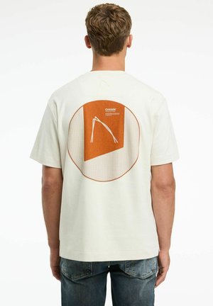 Beige katoenen t-shirt met korte mouwen, met een geometrisch oranje en witte ontwerp op de achterkant, en een gestructureerd cirkelpatroon.