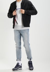 Zwarte bomberjack, grijze trui met rode accenten, lichtblauwe jeans, marineblauwe sneakers, geribbelde manchetten en een zakje met ritsdetail op het jack.