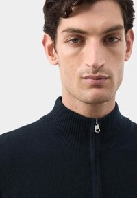 Felpa zip-up di un blu navy scuro con colletto a coste alto e una cerniera argentata, realizzata in un tessuto morbido. Inquadratura ravvicinata della parte superiore del corpo.