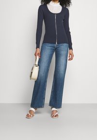 Marinblå ribbad tröja med dragkedja och silverdetaljer, tillsammans med vida blå jeans. En vit handväska fulländar outfiten.