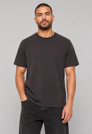 Mężczyzna z krótkimi włosami i brodą, ubrany w jednolity ciemnoszary t-shirt i czarne jeansy, stojący z jedną ręką w kieszeni na szarym tle.