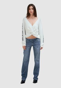 Weißer, flauschiger gestrickter Pullover mit einem verdrehten Vorderdesign und tiefem V-Ausschnitt, kombiniert mit hellblauen, geraden Jeans. Schwarze Akzente an den Ärmeln.
