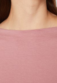 Světle růžový, měkký bavlněný top s širokým výstřihem, s hladkým povrchem a minimálním švem podél okrajů.