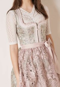 Vrouw in een getailleerde bloemencorset met roze kantaccenten over een witte kanten blouse, en een bijpassende roze kanten rok met een satijnen strikceintuur.