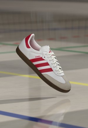 Λευκό αθλητικό παπούτσι Adidas Samba με κόκκινες ρίγες και σόλα από καουτσούκ που αιωρείται πάνω από το δάπεδο εσωτερικής αθλητικής αίθουσας με χρωματιστές γραμμές.